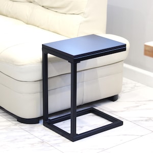 Pode incluir: Mesa de apoio de metal preto com tampo retangular e base em forma de C. A mesa está ao lado de um sofá branco.