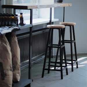 May include: Two black metal bar stools with round, light-coloured wooden seats. The stools are positioned in front of a wooden bar table. The table has a black metal frame and a light-coloured wooden top. The stools are approximately 76 cm tall.