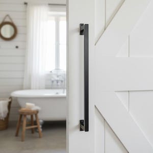 May include: A black, rectangular door pull is mounted on a white, paneled door. The door is part of a bathroom with a clawfoot tub, a small wooden stool, and a round mirror.