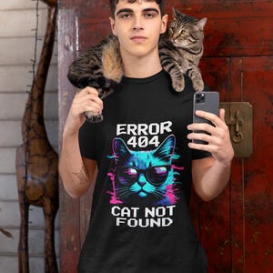 May include: Black t-shirt with a graphic of a cat wearing sunglasses and the text "ERROR 404 CAT NOT FOUND". The cat is rendered in shades of blue, pink, and white. A person is wearing the shirt and holding a phone.