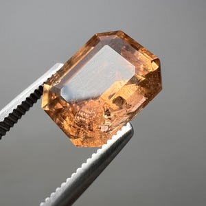 以下が含まれることがあります： 温かみのある琥珀色の長方形にカットされた宝石が、銀色のピンセットで挟まれています。 宝石の表面は光を反射し、その内部構造と透明度を示しています。 背景はニュートラルグレーで、宝石の色と形を際立たせています。