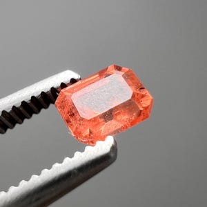 以下が含まれることがあります： オレンジがかった赤色の長方形の宝石が、銀色のピンセットで挟まれています。宝石は透明感のある内部を持ち、灰色の背景に配置されています。宝石のカットと色は、それが貴重な石であることを示唆しています。