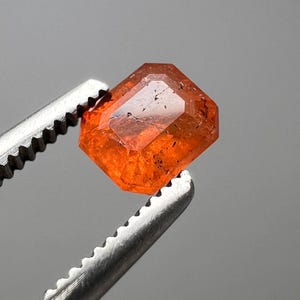 以下が含まれることがあります： 銀色のピンセットで保持された、ファセット加工されたオレンジ色の宝石。 宝石は、光を反射する複数のファセットを持つ八角形の形状をしています。 色は鮮やかなオレンジ色で、いくつかの暗いインクルージョンがあります。 背景はニュートラルグレーです。