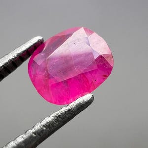 Może przedstawiać: Fasetowany, owalny kamień szlachetny rubinu, trzymany przez srebrną pęsetę. Klejnot ma żywy, głęboki różowy kolor z wewnętrznymi odbiciami światła. Pęseta jest wyraźnie ostra, podkreślając przejrzystość i blask klejnotu.