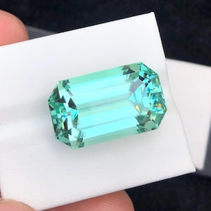 May include: A light blue, faceted, emerald-cut gemstone on a white background.
