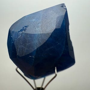 May include: A faceted, deep blue gemstone with a complex, geometric cut. The gem is mounted on a silver metal stand, showcasing its rich color and crystalline structure. The stone's facets reflect light, creating a play of shadows and highlights.