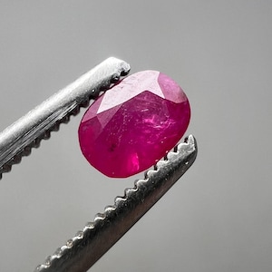 May include: A faceted, oval ruby gemstone held by silver tweezers. The ruby is a deep, vibrant red colour, with multiple facets catching the light. The tweezers have a serrated edge for gripping the stone.