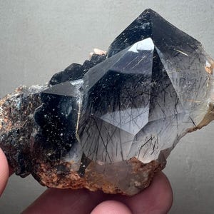 May include: A large, faceted quartz crystal with black tourmaline inclusions. The crystal is a deep, smoky grey colour with a complex, geometric shape. The base is a rough, brown rock.
