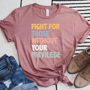 May include: Dusty rose t-shirt with the text "FIGHT FOR THOSE WITHOUT YOUR PRIVILEGE" in colorful block letters. The shirt is paired with denim shorts and brown suede shoes. The shirt is a casual wear item.