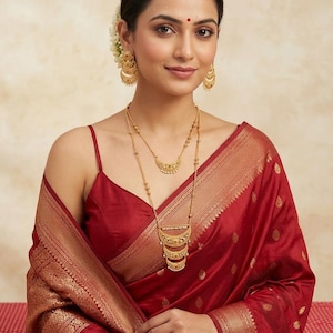 May include: A woman wearing a red sari with gold trim and a matching red blouse. She is adorned with gold earrings and a layered gold necklace. A flower is tucked into her hair. The sari is draped over her shoulder.