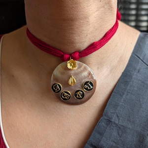 Puede incluir: Un collar con colgante redondo con un cordón rojo. El colgante es de color marrón claro con símbolos negros y detalles dorados. Una flor y una hoja doradas adornan la parte superior del colgante.