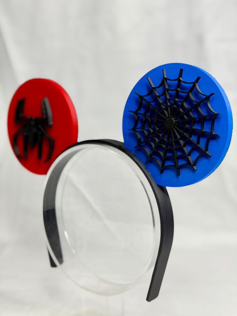 May include: Spider-Man themed mouse ears. The ears are red and blue circles with a black spider and spiderweb design. The headband is black with a clear plastic center.