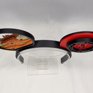 May include: A headband featuring two circular ears. The left ear displays a brown and tan desert landscape with small green accents. The right ear features a red and black wheel design with the text LIGHTYEAR printed in black on the outer rim.