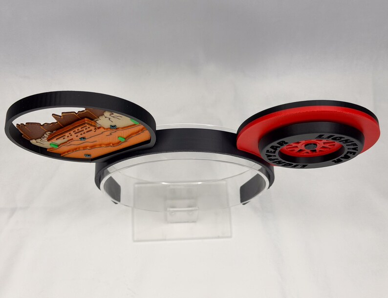 May include: A headband featuring two circular ears. The left ear displays a brown and tan desert landscape with small green accents. The right ear features a red and black wheel design with the text LIGHTYEAR printed in black on the outer rim.