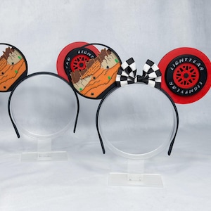 May include: Two headbands with circular ears. The left headband features ears with a desert landscape design. The right headband features ears with a black and red Lightyear tire design and a black and white checkered bow in the center.