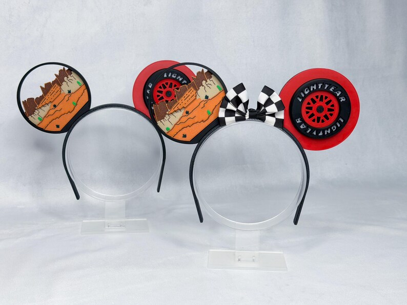 May include: Two headbands with circular ears. The left headband features ears with a desert landscape design. The right headband features ears with a black and red Lightyear tire design and a black and white checkered bow in the center.