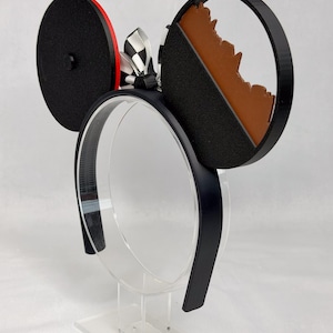 May include: A black headband featuring two circular ears. The left ear is solid black with a red side edge. The right ear is a black frame containing a brown, jagged mountain-like silhouette. A black and white checkered ribbon bow sits between the ears.