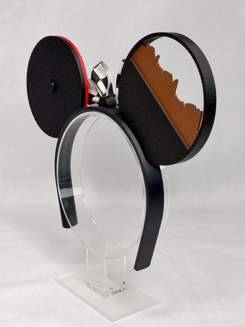 May include: A black headband featuring two circular ears. The left ear is solid black with a red side edge. The right ear is a black frame containing a brown, jagged mountain-like silhouette. A black and white checkered ribbon bow sits between the ears.