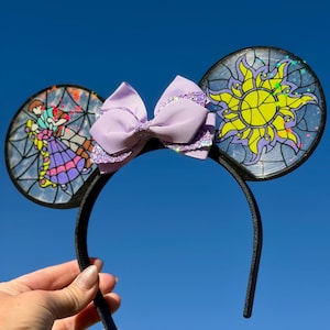 Puede incluir: Una diadema negra con dos orejas circulares con dise&ntilde;os estilo vitral. La oreja izquierda muestra a dos figuras bailando, mientras que la derecha exhibe un emblema de sol amarillo. Un lazo de cinta lavanda con detalles brillantes est&aacute; en el centro.