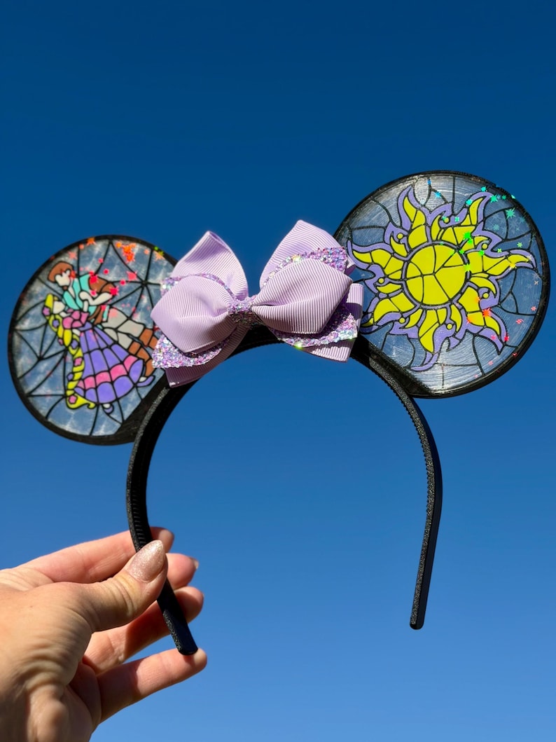 Puede incluir: Una diadema negra con dos orejas circulares con dise&ntilde;os estilo vitral. La oreja izquierda muestra a dos figuras bailando, mientras que la derecha exhibe un emblema de sol amarillo. Un lazo de cinta lavanda con detalles brillantes est&aacute; en el centro.