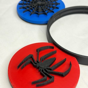 May include: A red, blue, and black spider-themed coaster set. The set includes a red coaster with a black spider, a blue coaster with a black spider web, and a black ring. The coasters are round and appear to be made of plastic.