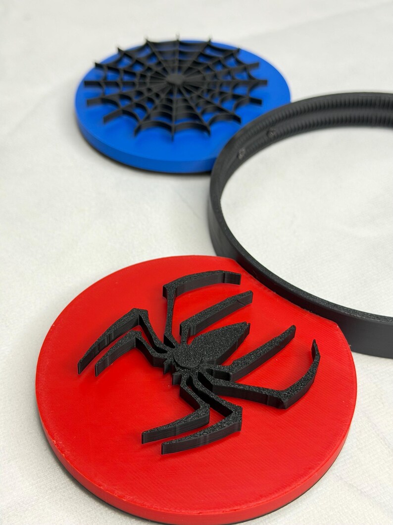May include: A red, blue, and black spider-themed coaster set. The set includes a red coaster with a black spider, a blue coaster with a black spider web, and a black ring. The coasters are round and appear to be made of plastic.