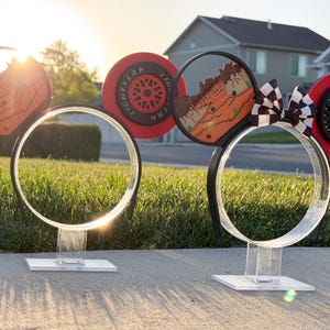 May include: Two sets of mouse-ear headbands displayed on clear acrylic stands. The designs feature red and black racing-themed elements, including tire-inspired circular motifs with Lightyear text, desert landscape graphics, and a black and white checkered bow.
