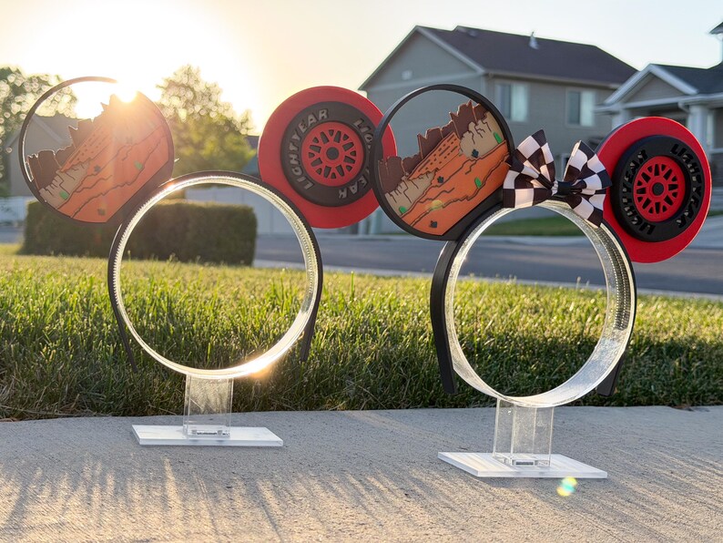May include: Two sets of mouse-ear headbands displayed on clear acrylic stands. The designs feature red and black racing-themed elements, including tire-inspired circular motifs with Lightyear text, desert landscape graphics, and a black and white checkered bow.