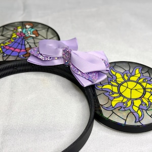Puede incluir: Una diadema negra con dos orejas circulares con dise&ntilde;os estilo vitral. Una oreja muestra a una pareja bailando, mientras que la otra presenta un emblema de sol amarillo. Un lazo de cinta morado claro superpuesto con detalles brillantes est&aacute; en el centro.