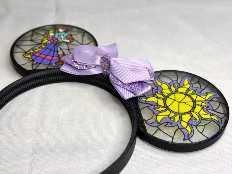 Puede incluir: Una diadema negra con dos orejas circulares con dise&ntilde;os estilo vitral. Una oreja muestra a una pareja bailando, mientras que la otra presenta un emblema de sol amarillo. Un lazo de cinta morado claro superpuesto con detalles brillantes est&aacute; en el centro.