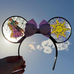 Puede incluir: Una diadema con dos orejas circulares con efecto de vidriera. La oreja izquierda muestra a una pareja bailando, mientras que la derecha presenta un emblema de sol amarillo. Un lazo morado claro con detalles brillantes se sit&uacute;a entre las orejas.