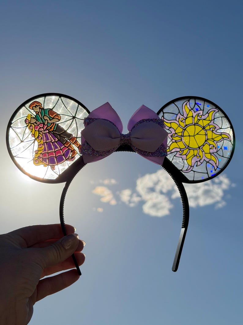 Puede incluir: Una diadema con dos orejas circulares con efecto de vidriera. La oreja izquierda muestra a una pareja bailando, mientras que la derecha presenta un emblema de sol amarillo. Un lazo morado claro con detalles brillantes se sit&uacute;a entre las orejas.