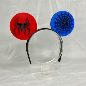May include: A black headband with two round, decorative elements. One is red with a black spider, the other is blue with a black spider web design. The headband is displayed on a clear stand.