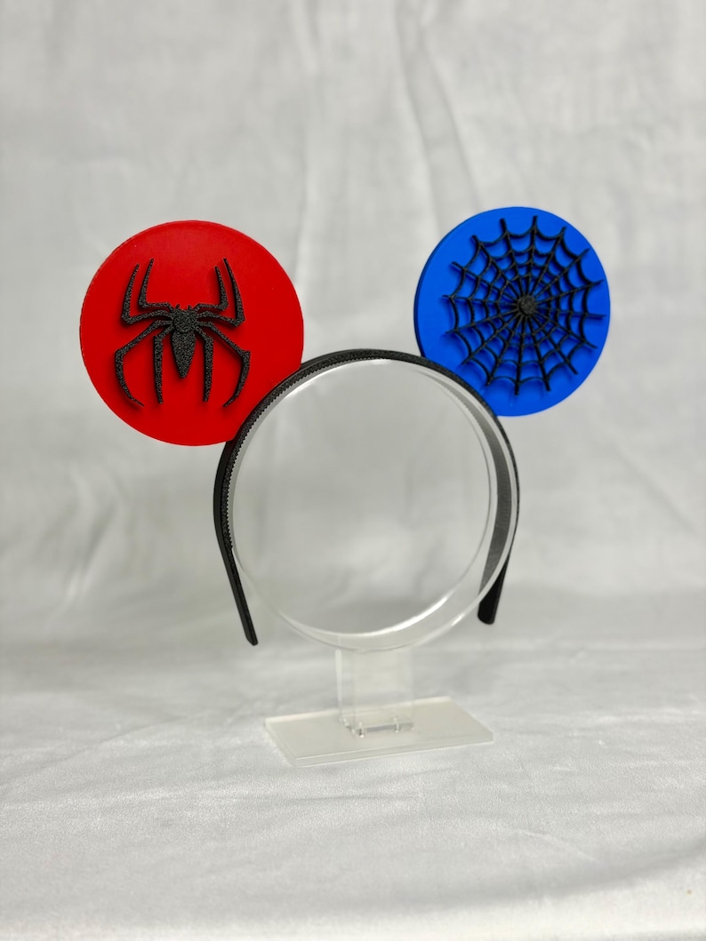 May include: A black headband with two round, decorative elements. One is red with a black spider, the other is blue with a black spider web design. The headband is displayed on a clear stand.