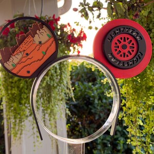 May include: A headband featuring two circular ears. The left ear displays a desert landscape with orange rock formations. The right ear is a red and black wheel design with the text LIGHTYEAR embossed in black lettering along the outer rim.