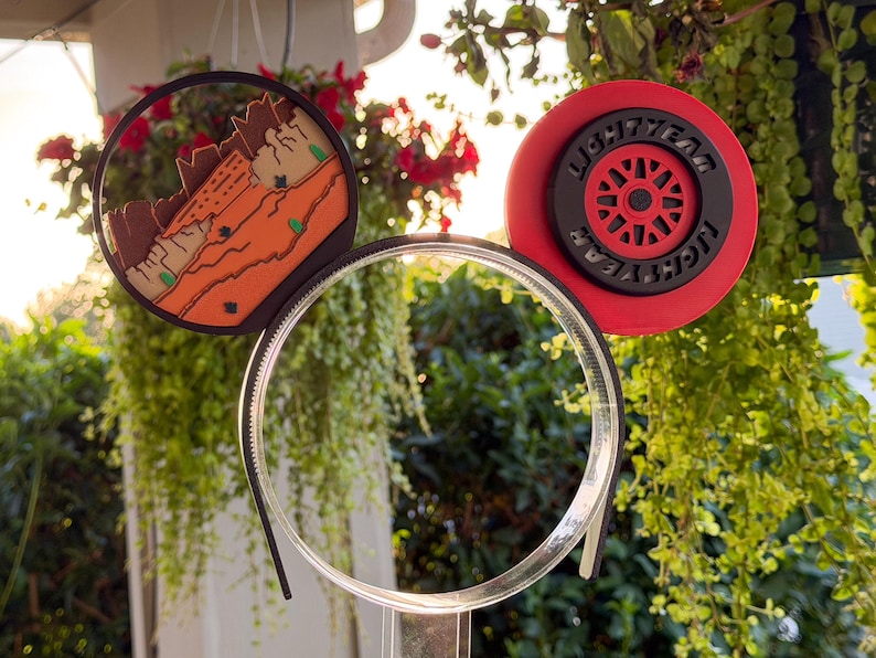 May include: A headband featuring two circular ears. The left ear displays a desert landscape with orange rock formations. The right ear is a red and black wheel design with the text LIGHTYEAR embossed in black lettering along the outer rim.