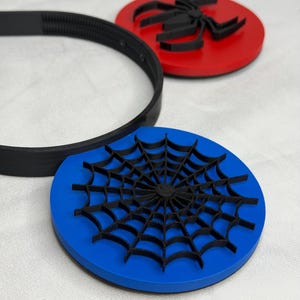 May include: A black headband and two round coasters. One coaster is red with a black spider design. The other coaster is blue with a black spider web design. The items are on a white surface.