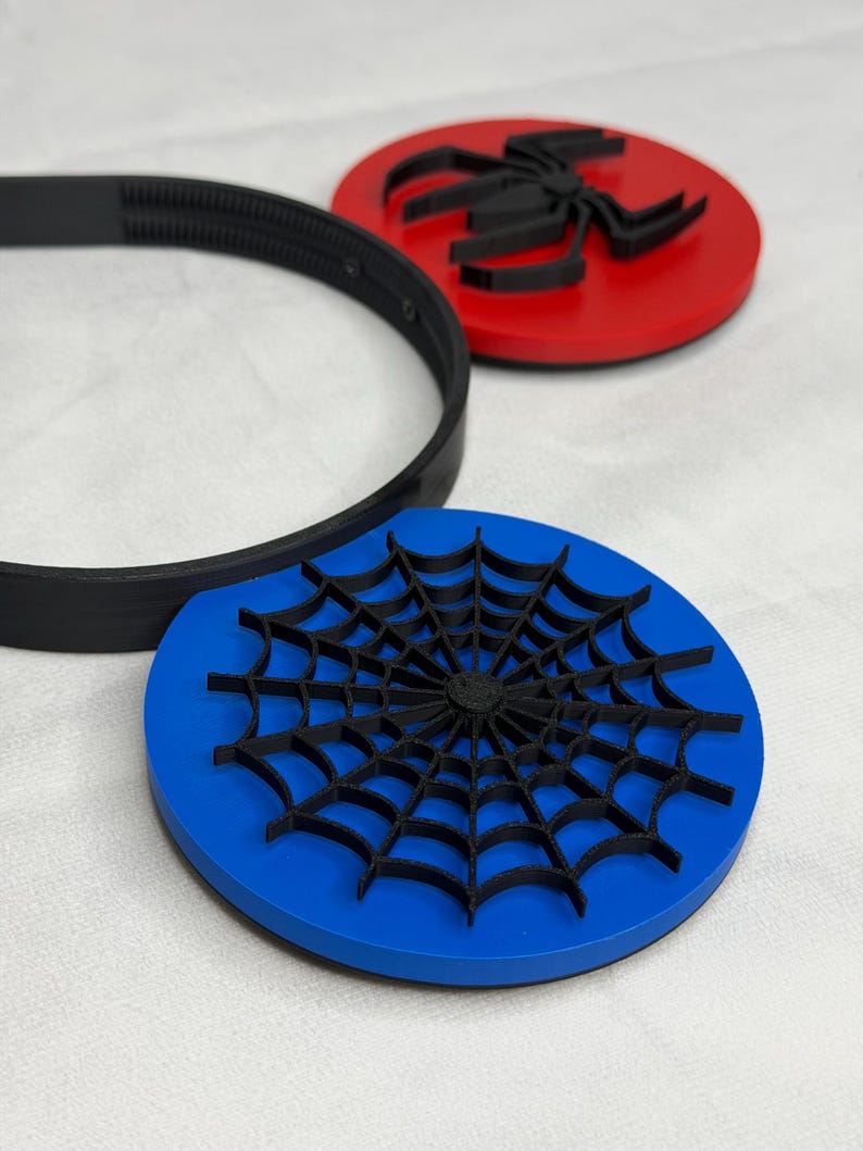 May include: A black headband and two round coasters. One coaster is red with a black spider design. The other coaster is blue with a black spider web design. The items are on a white surface.