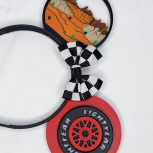 May include: A black headband featuring two circular ears. One ear displays a desert landscape with orange rock formations. The other ear features a red tire with the text Lightyear. A black and white checkered bow sits between the two ears.