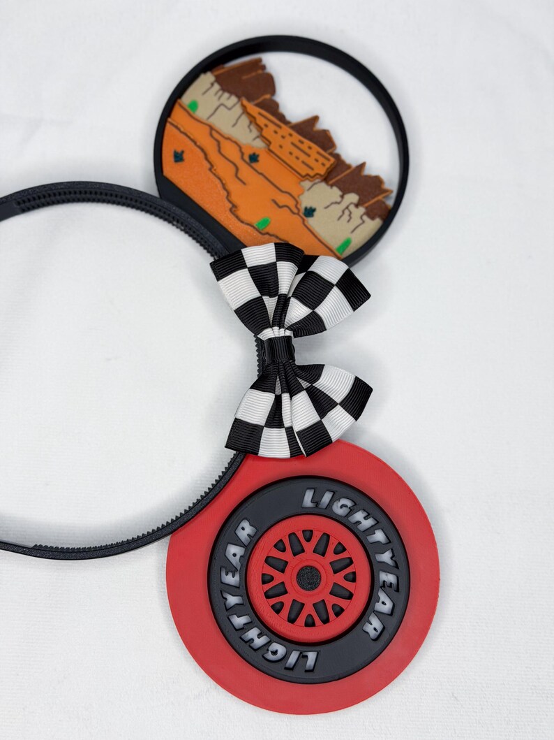 May include: A black headband featuring two circular ears. One ear displays a desert landscape with orange rock formations. The other ear features a red tire with the text Lightyear. A black and white checkered bow sits between the two ears.