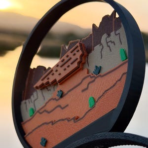 May include: A circular black frame containing a layered desert landscape relief. The scene features textured brown and tan rock formations, orange plateaus, and small green cacti accents. The design is mounted on a black headband base.