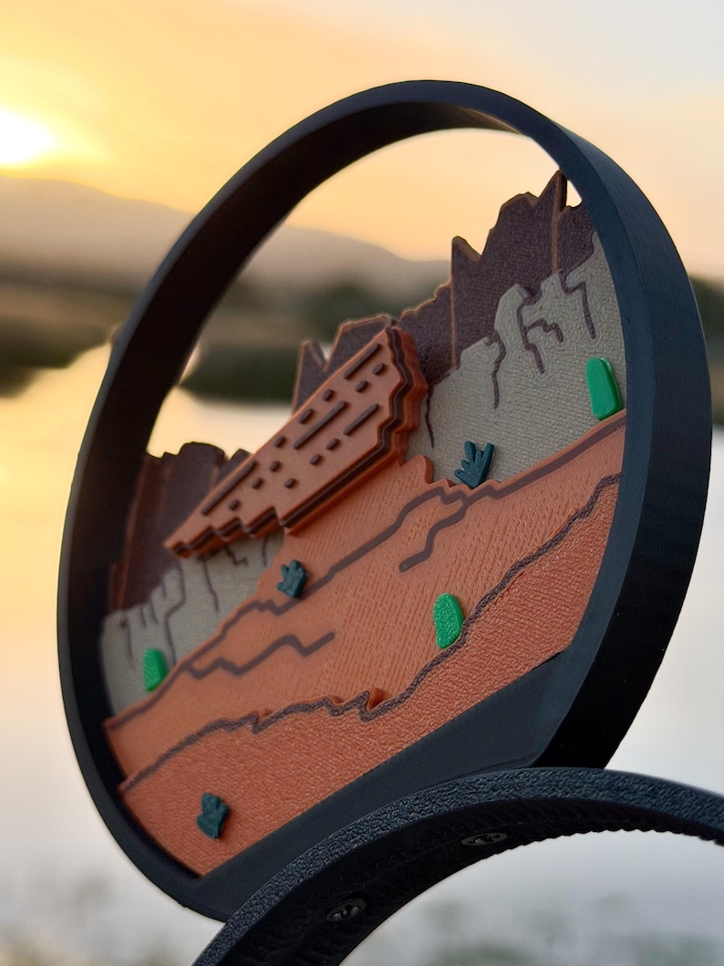 May include: A circular black frame containing a layered desert landscape relief. The scene features textured brown and tan rock formations, orange plateaus, and small green cacti accents. The design is mounted on a black headband base.