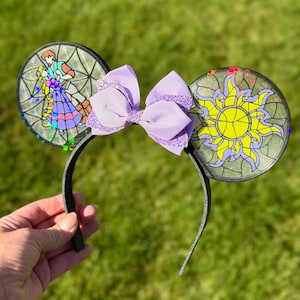 Puede incluir: Una diadema con dos orejas circulares con efecto de vidriera. La oreja izquierda representa a una pareja bailando, mientras que la derecha muestra un emblema de sol amarillo. Un lazo superpuesto de color morado claro con purpurina se sit&uacute;a entre las orejas.
