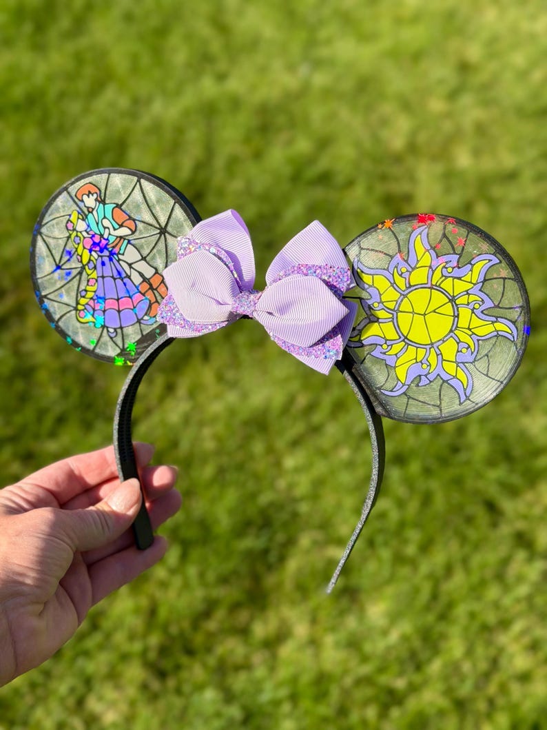 Puede incluir: Una diadema con dos orejas circulares con efecto de vidriera. La oreja izquierda representa a una pareja bailando, mientras que la derecha muestra un emblema de sol amarillo. Un lazo superpuesto de color morado claro con purpurina se sit&uacute;a entre las orejas.