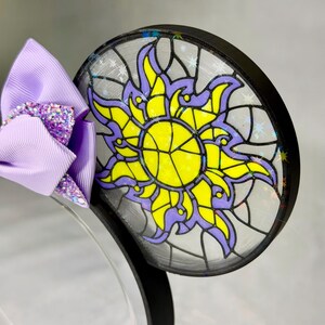 Puede incluir: Una diadema con una oreja circular que presenta un dise&ntilde;o de sol estilo vidriera en amarillo y morado, acentuado con detalles de estrellas hologr&aacute;ficas. Un lazo de cinta lavanda con un centro de purpurina morada est&aacute; unido al lado.