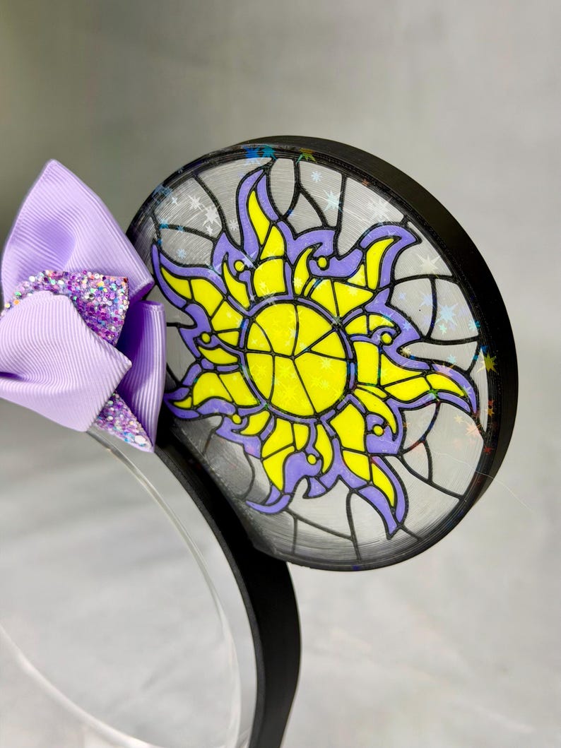 Puede incluir: Una diadema con una oreja circular que presenta un dise&ntilde;o de sol estilo vidriera en amarillo y morado, acentuado con detalles de estrellas hologr&aacute;ficas. Un lazo de cinta lavanda con un centro de purpurina morada est&aacute; unido al lado.