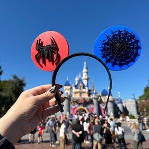 May include: Black headband with red and blue Mickey Mouse ears. The red ear features a black spider, and the blue ear has a black spiderweb design. The background includes a castle and a crowd of people.