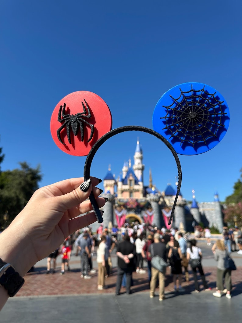 May include: Black headband with red and blue Mickey Mouse ears. The red ear features a black spider, and the blue ear has a black spiderweb design. The background includes a castle and a crowd of people.