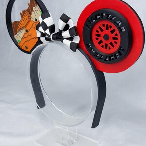 May include: A headband featuring two circular ears. The left ear displays a desert landscape with orange rock formations. The right ear features a red and black wheel design with the text LIGHTYEAR embossed around the center. A black and white checkered bow sits between the ears.