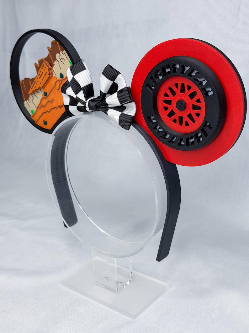 May include: A headband featuring two circular ears. The left ear displays a desert landscape with orange rock formations. The right ear features a red and black wheel design with the text LIGHTYEAR embossed around the center. A black and white checkered bow sits between the ears.